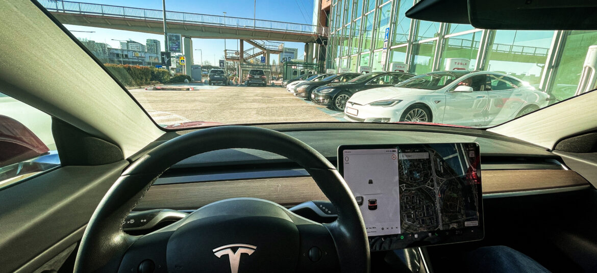 Pohlad z Tesly, z pohladu vodiča/vodičky ponad volant s logom Tesla na Tesla Supercharger pri Auparku v Bratislave plane obsadeny nabijajucimi vozidlami Tesla..